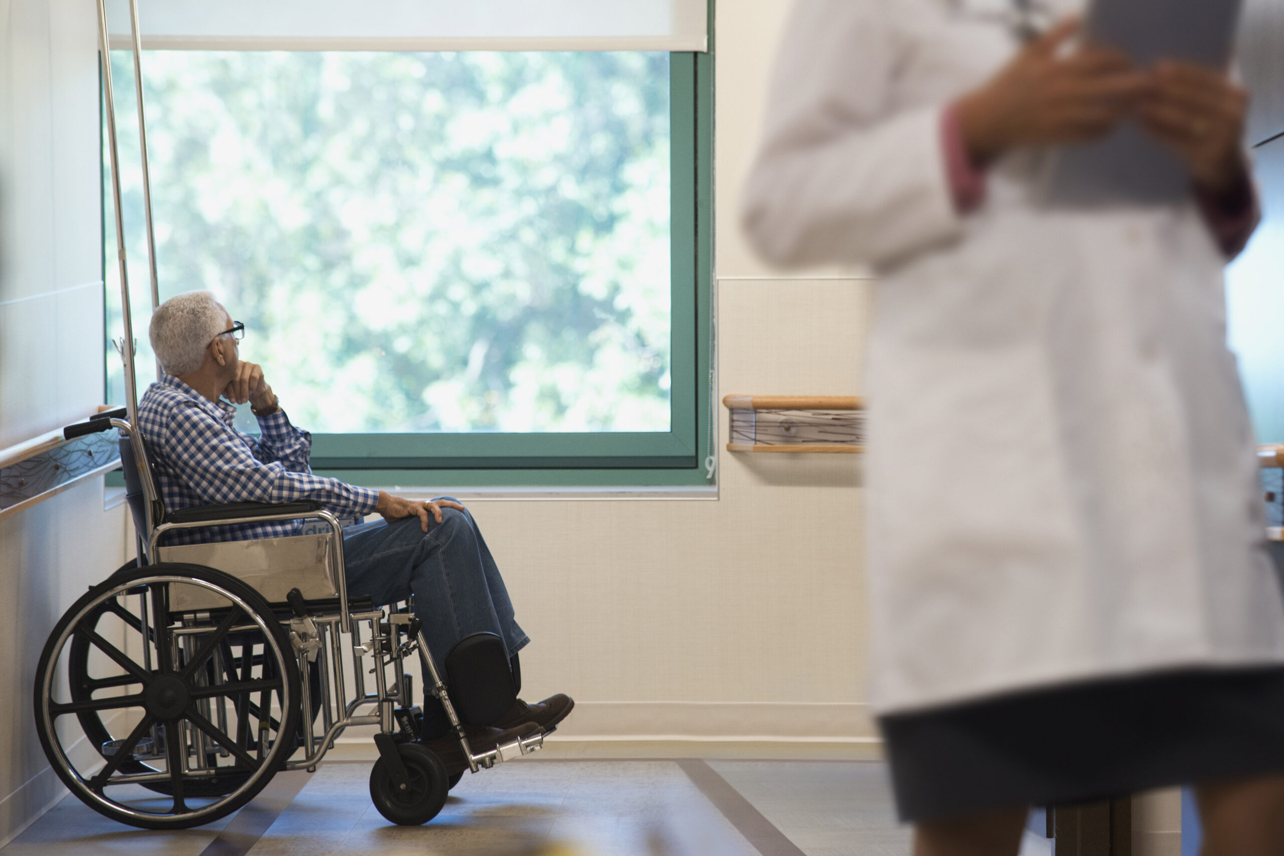 An elderly man is sitting in a wheelchair in a hospital room. A health care worker is visible in the foreground of the image.