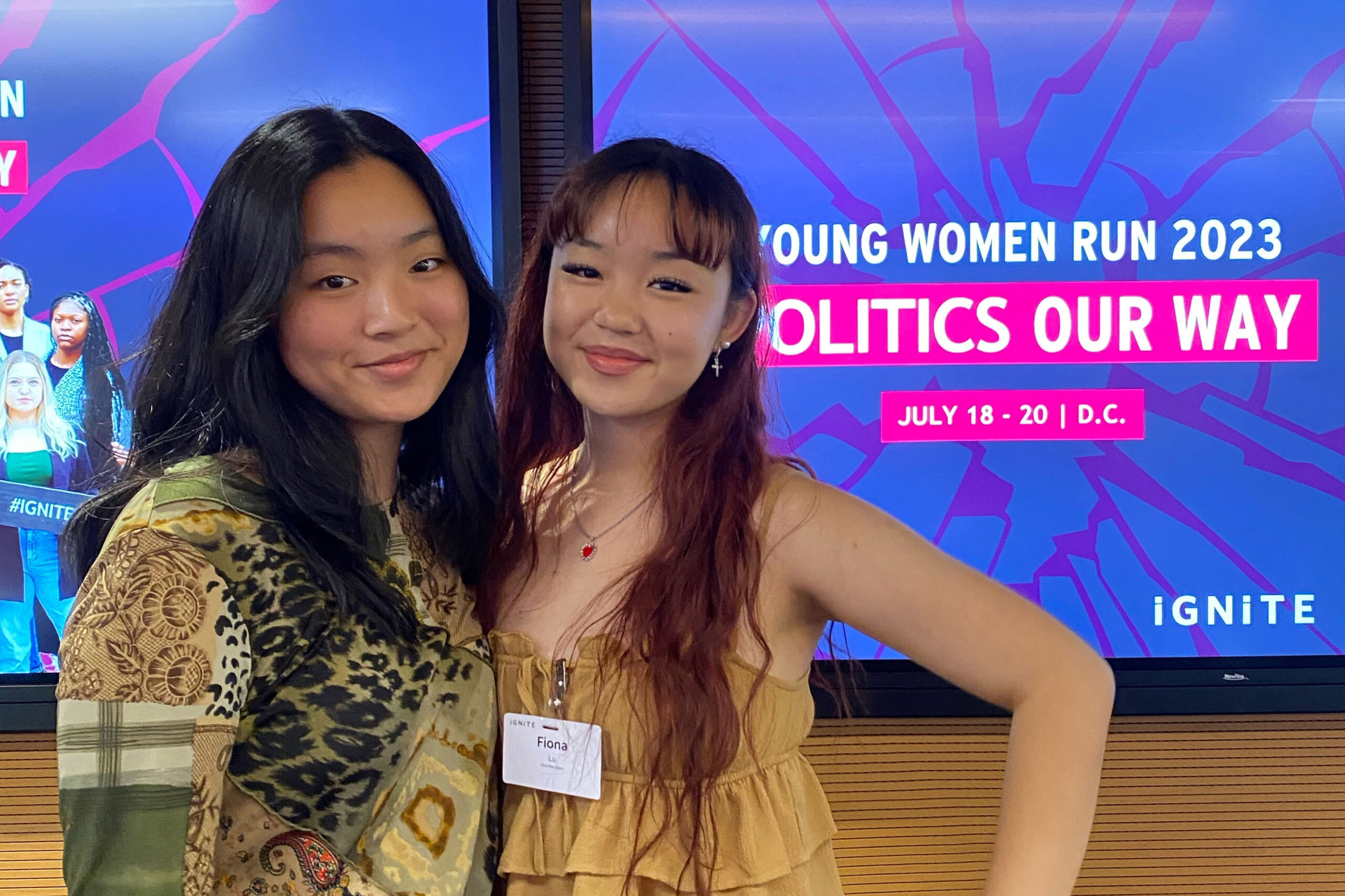 A photo of two teenagers posing for a photo together. They stand in front of a screen that reads, 